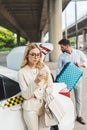 young blonde woman in eyeglasses using smartphone while man putting suitcase in trunk of taxi Royalty Free Stock Photo