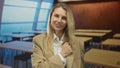 Young blonde woman gesturing confidently in an empty modern classroom setting with tables and large windows in the background Royalty Free Stock Photo