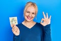 Young blonde woman holding anarchism drawing reminder doing ok sign with fingers, smiling friendly gesturing excellent symbol