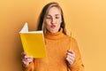 Young blonde woman holding book and pencil making fish face with mouth and squinting eyes, crazy and comical