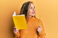 Young blonde woman holding book and pencil smiling looking to the side and staring away thinking Royalty Free Stock Photo