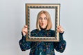 Young blonde woman holding empty frame making fish face with mouth and squinting eyes, crazy and comical Royalty Free Stock Photo