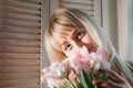 A young blonde woman holding flowers by the window, smiling Royalty Free Stock Photo