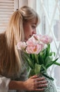 Young blonde woman holding flowers by the window, smiling Royalty Free Stock Photo