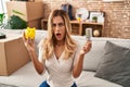 Young blonde woman holding led bulb and piggy bank in shock face, looking skeptical and sarcastic, surprised with open mouth Royalty Free Stock Photo