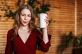 Young blonde woman holding a paper cup of hot coffee of takeaway in her hands indoors in cafe and looking in camera. copyspace. Royalty Free Stock Photo