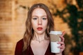 Young blonde woman holding a paper cup of hot coffee of takeaway in her hands indoors in cafe and looking in camera. copyspace. Royalty Free Stock Photo