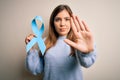 Young blonde woman holding prostate cancer awareness campaing blue ribbon with open hand doing stop sign with serious and Royalty Free Stock Photo