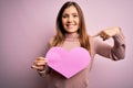 Young blonde woman holding romantic big paper heart shape over pink isolated background with surprise face pointing finger to Royalty Free Stock Photo