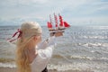 Young blonde woman holding small yacht with red sails on sea beach