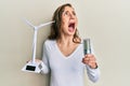 Young blonde woman holding solar windmill for renewable electricity and led light bulb angry and mad screaming frustrated and