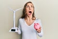 Young blonde woman holding solar windmill for renewable electricity and piggy bank angry and mad screaming frustrated and furious,