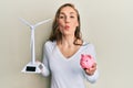 Young blonde woman holding solar windmill for renewable electricity and piggy bank looking at the camera blowing a kiss being Royalty Free Stock Photo