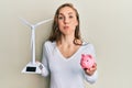 Young blonde woman holding solar windmill for renewable electricity and piggy bank puffing cheeks with funny face