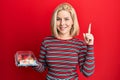 Young blonde woman holding take away food smiling with an idea or question pointing finger with happy face, number one