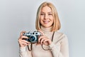 Young blonde woman holding vintage camera looking positive and happy standing and smiling with a confident smile showing teeth Royalty Free Stock Photo