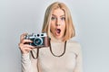 Young blonde woman holding vintage camera scared and amazed with open mouth for surprise, disbelief face Royalty Free Stock Photo