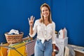 Young blonde woman at laundry room showing and pointing up with fingers number four while smiling confident and happy