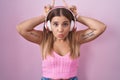 Young blonde woman listening to music using headphones doing funny gesture with finger over head as bull horns