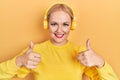 Young blonde woman listening to music using headphones success sign doing positive gesture with hand, thumbs up smiling and happy Royalty Free Stock Photo