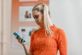 A young blonde woman with long hair dressed in an orange sweater stands and holds a phone in her hands and looks at the phone scre