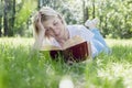 A young blonde woman is lying on the grass in a park on a sunny summer day and is reading a book. Recreation and distance learning Royalty Free Stock Photo
