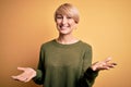 Young blonde woman with modern short hair wearing casual sweater over yellow background smiling cheerful with open arms as Royalty Free Stock Photo