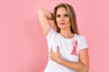 Young blonde woman with pink ribbon on t shirt check her breast with hand on pink background. Breast cancer awareness.