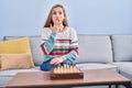 Young blonde woman playing chess sitting on the sofa covering mouth with hand, shocked and afraid for mistake Royalty Free Stock Photo