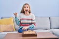 Young blonde woman playing chess sitting on the sofa pointing thumb up to the side smiling happy with open mouth Royalty Free Stock Photo