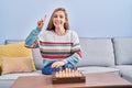 Young blonde woman playing chess sitting on the sofa smiling with an idea or question pointing finger with happy face, number one Royalty Free Stock Photo