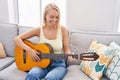 Young blonde woman playing classical guitar sitting on sofa at home Royalty Free Stock Photo