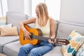 Young blonde woman playing classical guitar sitting on sofa at home Royalty Free Stock Photo