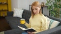 Young blonde woman reading book and drinking coffee sitting on sofa at home Royalty Free Stock Photo