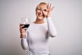 Young blonde woman with short hair drinking a glass of red wine over isolated background with happy face smiling doing ok sign Royalty Free Stock Photo