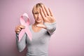 Young blonde woman with short hair holding breast cancer awareness pink ribbon with open hand doing stop sign with serious and Royalty Free Stock Photo