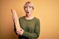 Young blonde woman with short hair holding fresh bread baguette over yellow background scared in shock with a surprise face,