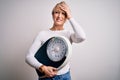 Young blonde woman with short hair holding scale for healthy weight and lifestyle stressed with hand on head, shocked with shame