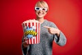 Young blonde woman with short hair wearing 3d movie glasses and eating popcorn at the cinema very happy pointing with hand and Royalty Free Stock Photo