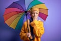 Young blonde woman with short hair wearing waterproff raincoat and colorful umbrella cover mouth with hand shocked with shame for