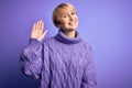 Young blonde woman with short hair wearing winter turtleneck sweater over purple background Waiving saying hello happy and