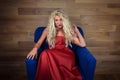 Young blonde woman sits on armchair. Elegant girl in red dress posing on camera Royalty Free Stock Photo