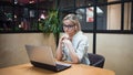 Young blonde woman sits at her laptop and uses wireless headphones.