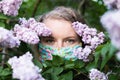 Young blonde woman smells lilac tree blossom through facial mask. Conceptual image of corona virus quarantine Royalty Free Stock Photo