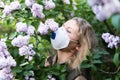 Young blonde woman smells lilac tree blossom through FFP2 respirator. Conceptual image of corona virus quarantine Royalty Free Stock Photo