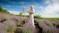 Young blonde woman standing in lavender field and making painting on easel