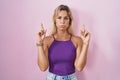 Young blonde woman standing over pink background pointing up looking sad and upset, indicating direction with fingers, unhappy and Royalty Free Stock Photo