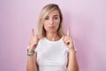 Young blonde woman standing over pink background pointing up looking sad and upset, indicating direction with fingers, unhappy and Royalty Free Stock Photo