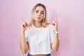 Young blonde woman standing over pink background pointing up looking sad and upset, indicating direction with fingers, unhappy and Royalty Free Stock Photo
