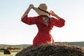 Blonde woman in hat and red dress is sitting on a haystack. View from the back